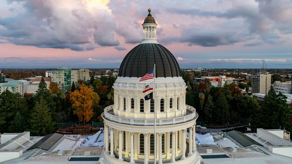 The dome of the California State Capitol building in Sacramento, Calif., with U.S. and California flags flying at sunset against a colorful sky