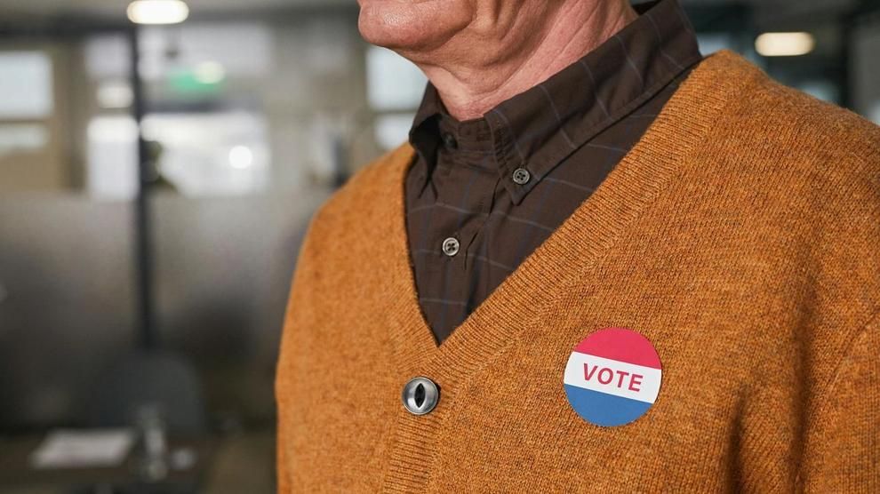 A man wearing a brown sweater and plaid shirt has a round red, white, and blue “VOTE” button pinned to his chest. Photo: Edmond Dantès / Pexels