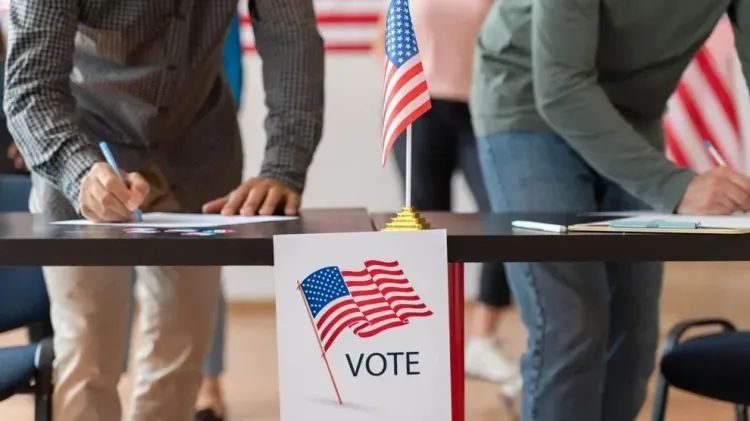 Voters fill out registration forms at a table decorated with an American flag and a “VOTE” sign inside a polling location.