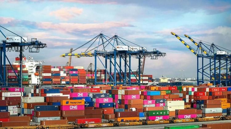 Colorful shipping containers are stacked beneath large blue cranes at the Port of Los Angeles in California, with a cargo ship docked in the background.