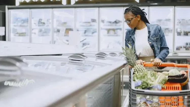 A woman pushes a shopping cart filled with fresh produce while browsing the frozen food aisle at a supermarket.