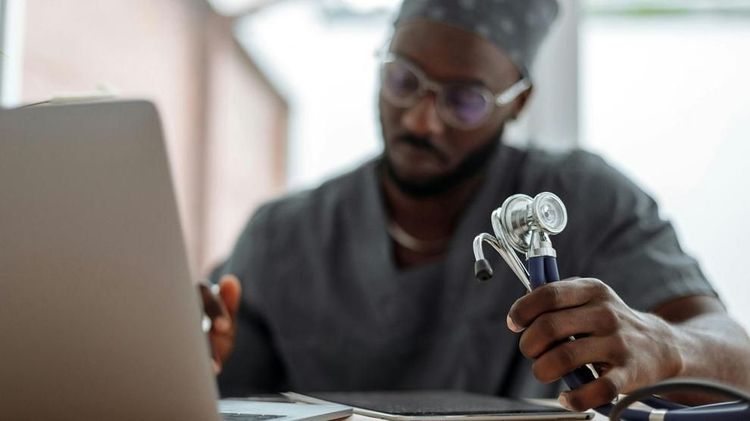 A physician in scrubs reviews patient data on a laptop while holding a stethoscope. Photo: Thirdman / Pexels