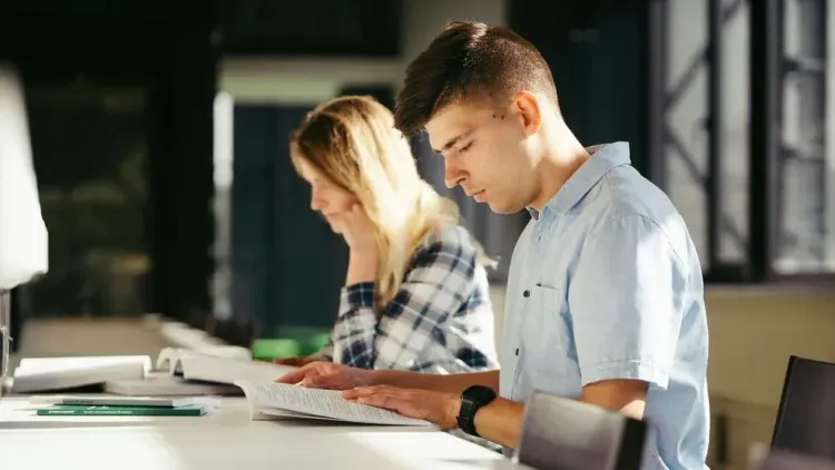 Two college students read textbooks at a shared study table.