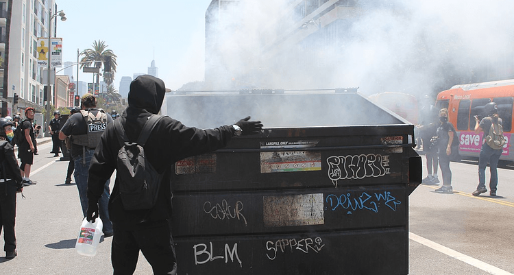 An ANTIFA activist holding a gallon of water inspects a smoldering dumpster lit on fire by other activists. Around the sides of the dumpster can be seen other activists and press individuals, some taking pictures, some watching.