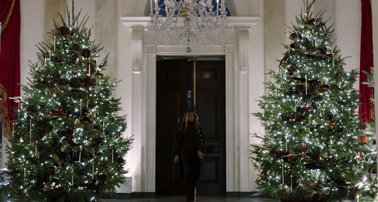 Melanie Trump walking toward the camera between two classically decorated Christmas trees.