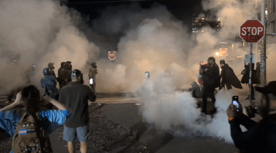 Protestors seen facing off with federal Agents in riot gear as smoke swirls.